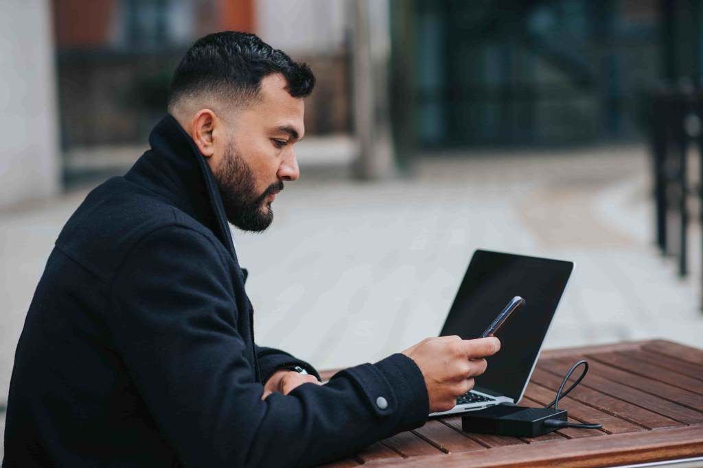  homem usando um casaco de frio preto utilizando um celular na mão direta e também um notebook na mesa em sua frente em referência ao aprendizado sobre hedge