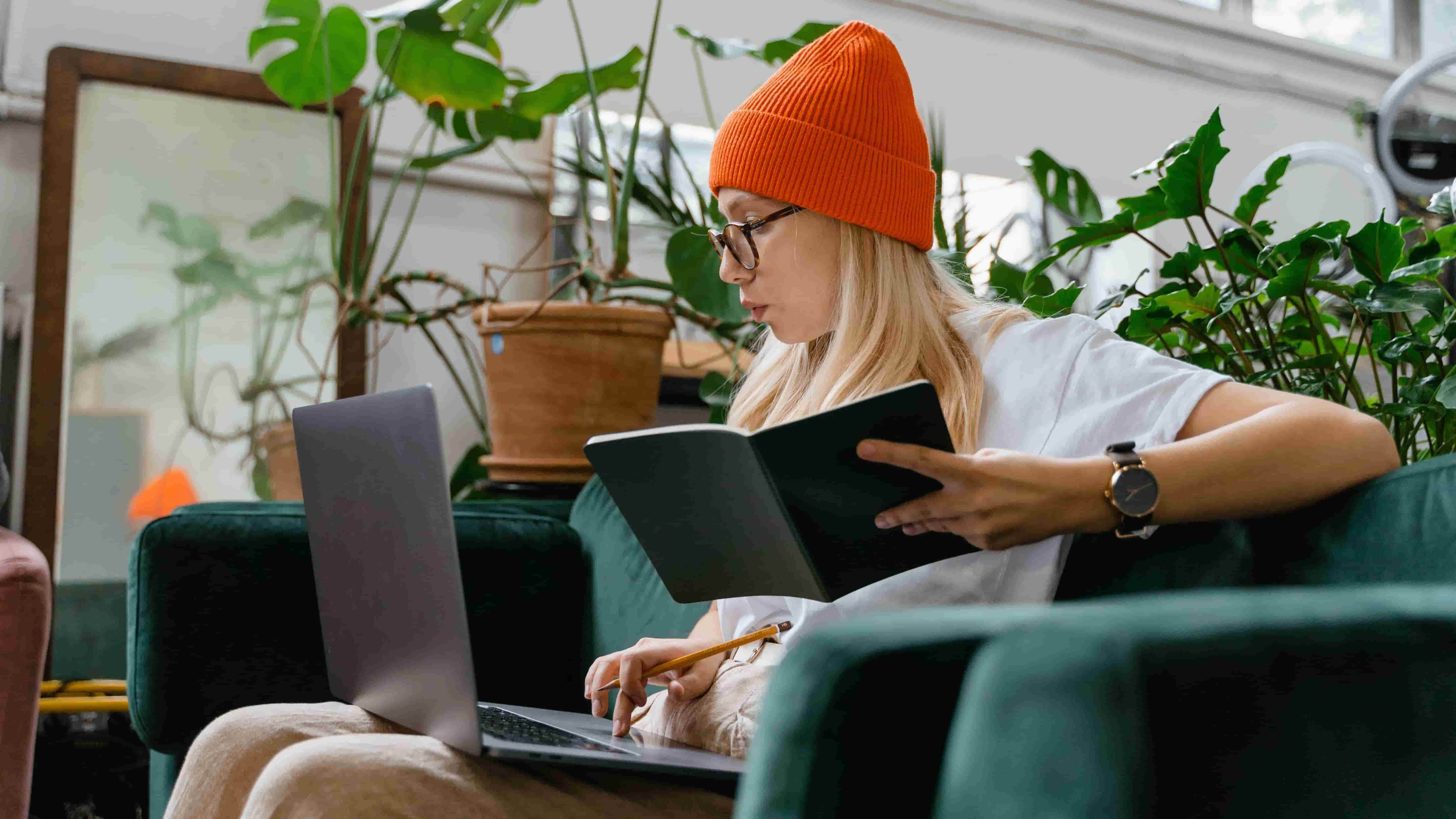 mulher sentada em um sofá da cor verde. Ela é branca e loira e está vestindo uma camiseta branca e uma touca laranja. Ela está usando um notebook em seu colo e está segurança um caderno na outra mão em referência a definição de metas para sua carteira de investimentos.