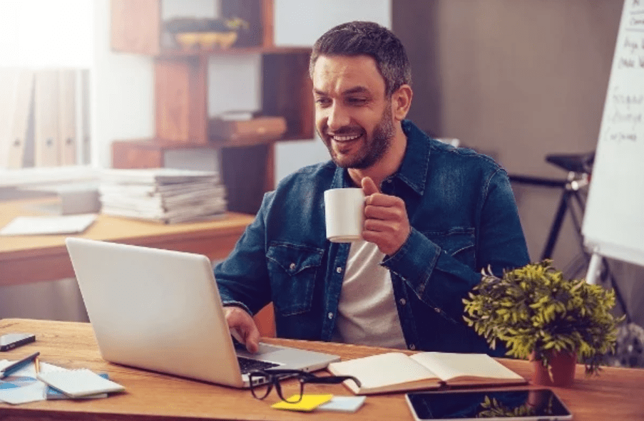 Um homem segurando uma xícara e olhando para o notebook feliz, em referência a como poupar dinheiro.