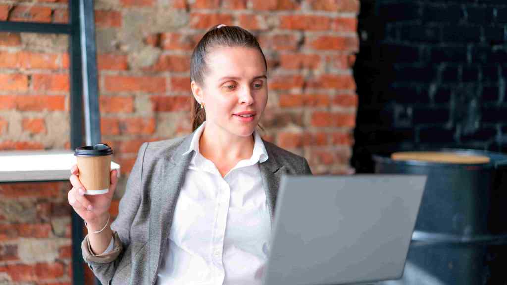 mulher segurando um café na mão direta e utilizando um notebook com a outra mão relacionado às pesquisas sobre investimentos para iniciantes