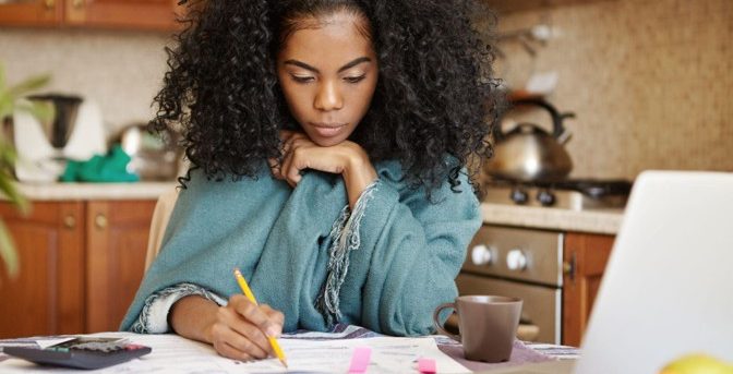 Mulher concentrada anotando gastos e contas a pagar em uma mesa com notebook, calculadora e papéis organizados para montar seu planejamento financeiro pessoal.