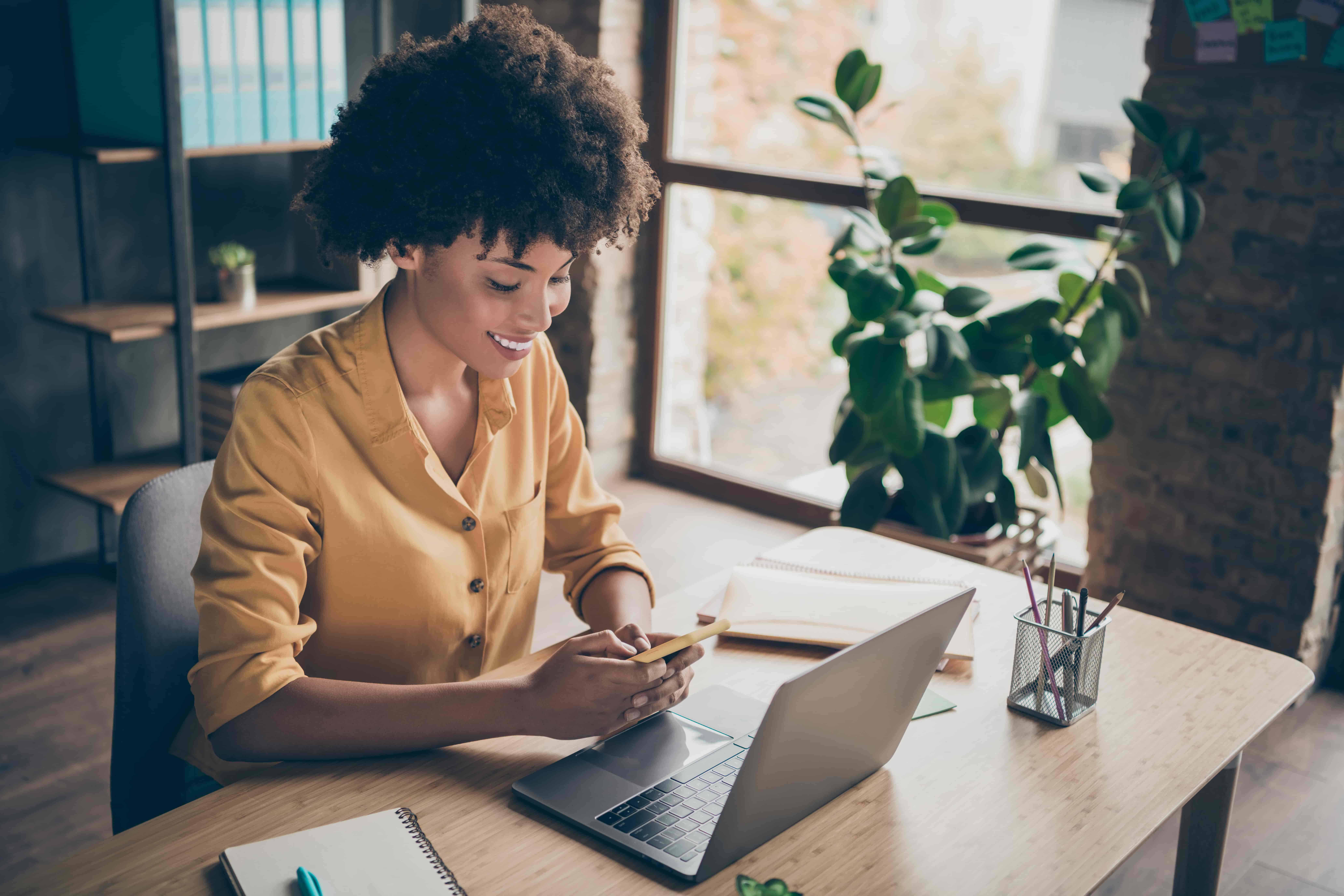 A imagem mostra uma mulher negra de camisa amarela utilizando um celular e um notebook em uma mesa de escritório em referência ao Tesouro Direto no Imposto de Renda.