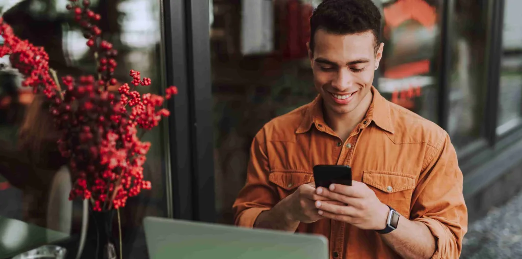 A imagem mostra um homem sorrindo segurando um celular. Ele está sentado em uma mesa com um notebook em referência ao processo de descoberta dos investimentos sem Imposto de Renda.