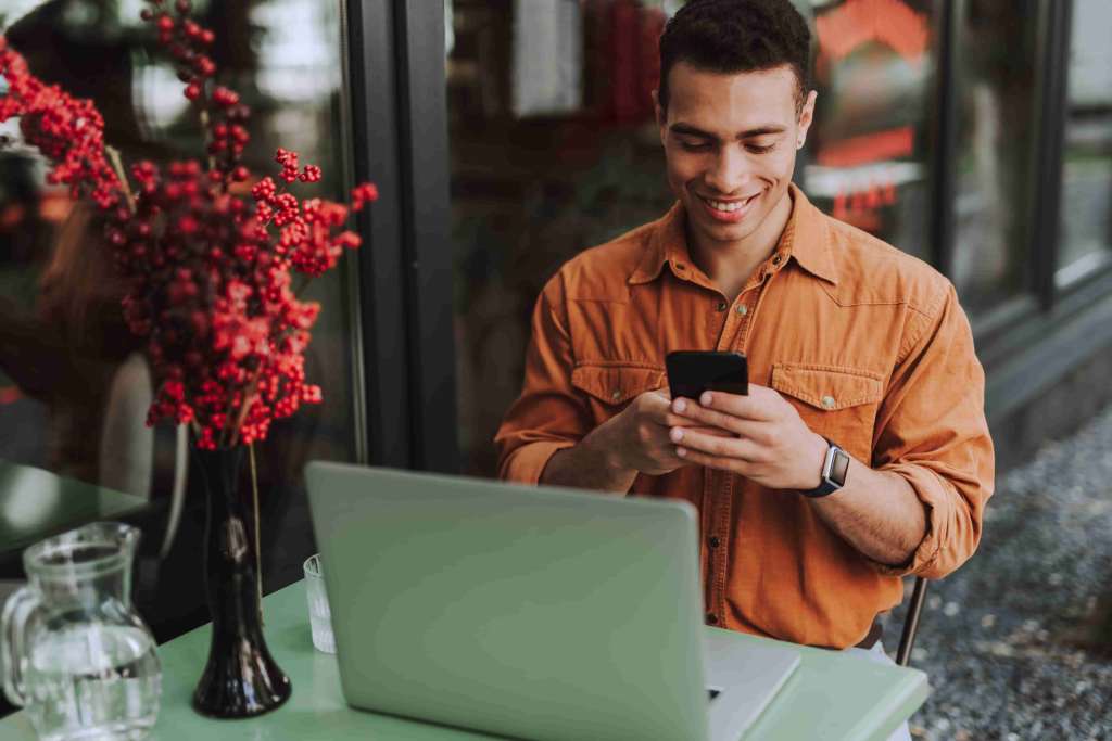 A imagem mostra um homem sorrindo segurando um celular. Ele está sentado em uma mesa com um notebook em referência ao processo de descoberta dos investimentos sem Imposto de Renda.