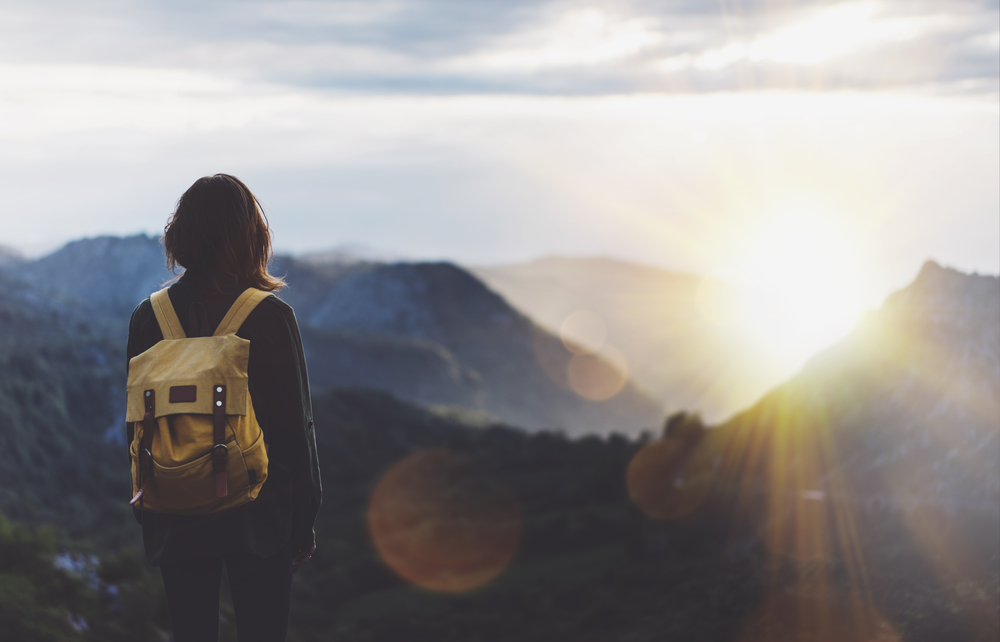 Uma mulher está de costas para a câmera enquanto observa o pôr do sol nas montanhas, em referência a como juntar dinheiro para viajar.