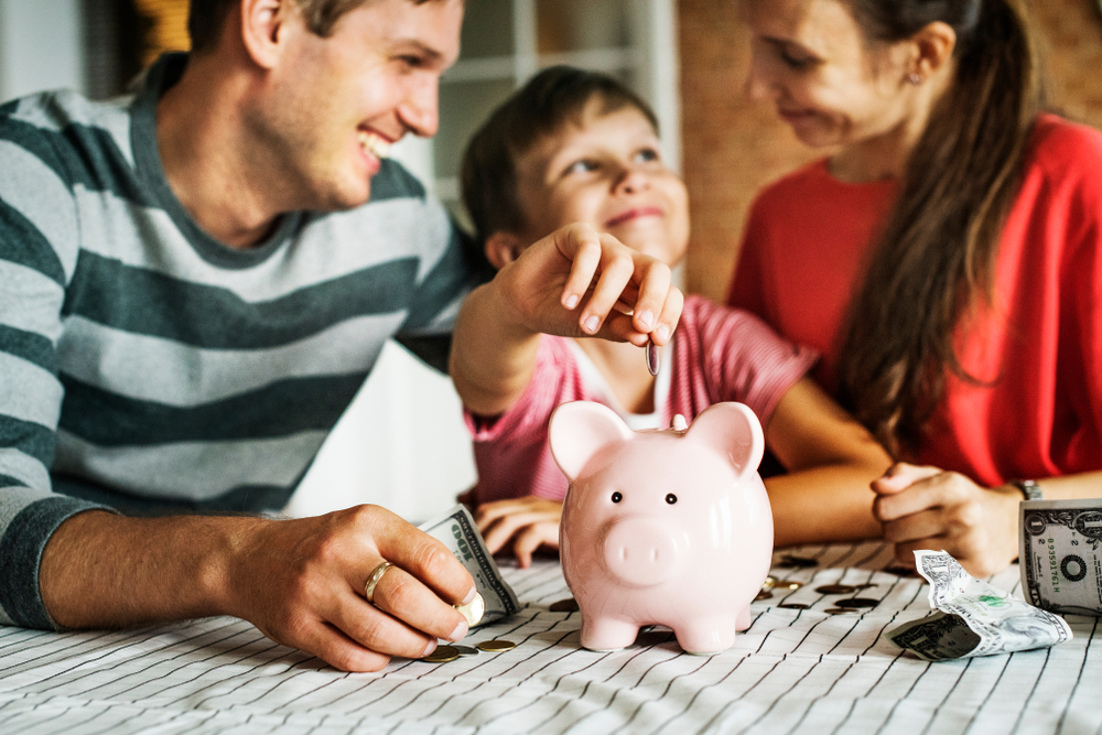 Uma família debruçada sobre a cama. O pai e a mãe olham par a criança sorridentes, enquanto ela coloca uma moeda no cofrinho, em referência a como se organizar financeiramente.