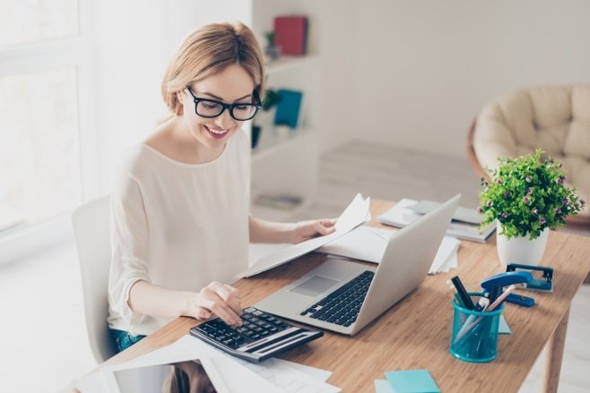 Uma moça sentada à mesa, que contém um notebook. Ela mexe na calculadora, em referência à dúvida sobre corretora de valores e bancos