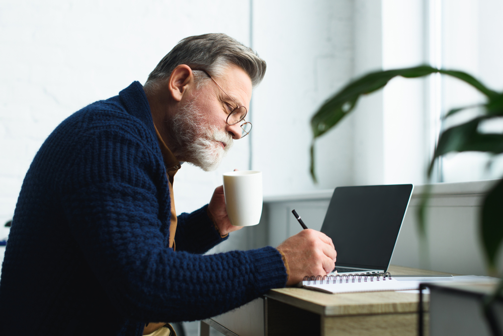 Um homem mais velho à mesa, com uma xícara na mão faz anotações em um caderno, em referência ao inss ou previdência privada