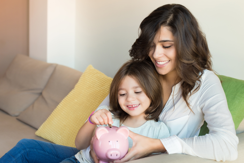 Uma mão e uma filha sentadas no sofá. A filha coloca uma moeda em um cofrinho em referência aos melhores investimentos para o futuro do meu filho