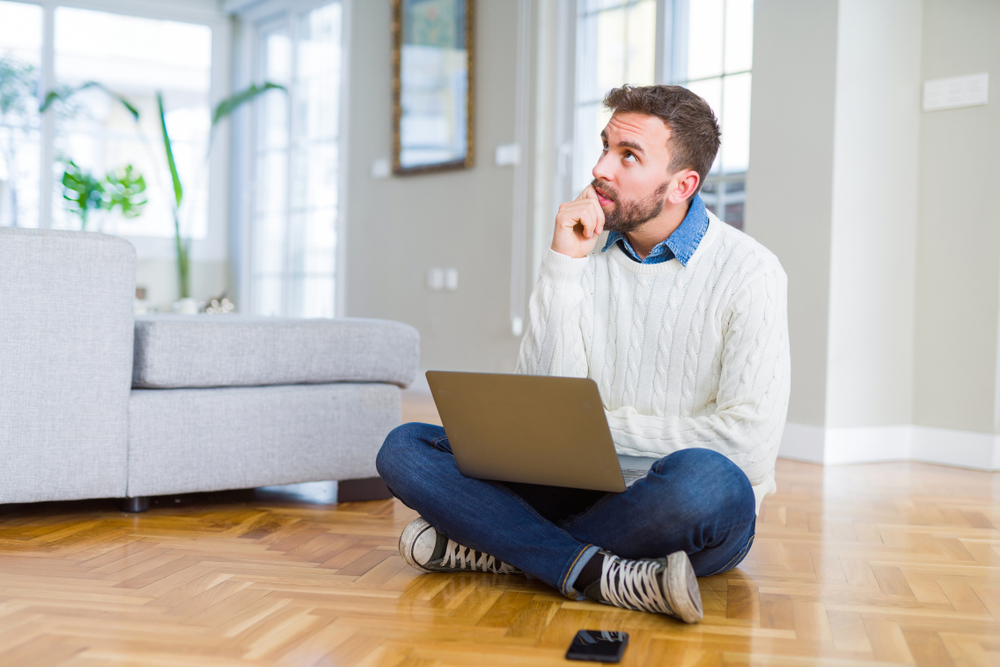 Um homem sentado de pernas cruzadas no chão olhando para cima com expressão de pensamento. No colo, um notebook, em referência à dúvida sobre viver de dividendos