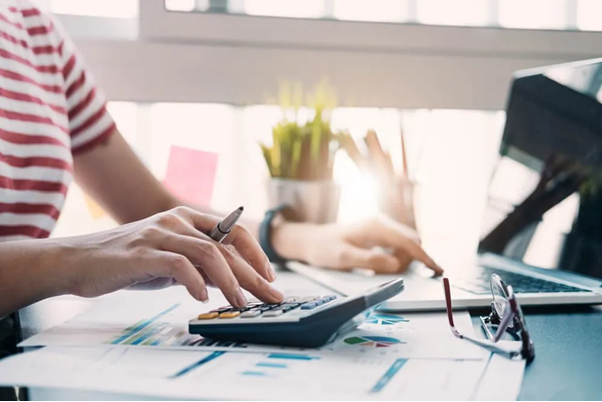 Mão feminina utiliza uma calculadora enquanto a outra mão digita no notebook, ambas posicionadas sobre uma mesa de trabalho. No ambiente, há também um óculos de grau e documentos com gráficos financeiros espalhados, sugerindo um cenário de análise detalhada. A imagem simboliza o cálculo do ponto de equilíbrio contábil, destacando a importância do uso de ferramentas e da análise de dados para a gestão financeira eficiente de uma empresa.