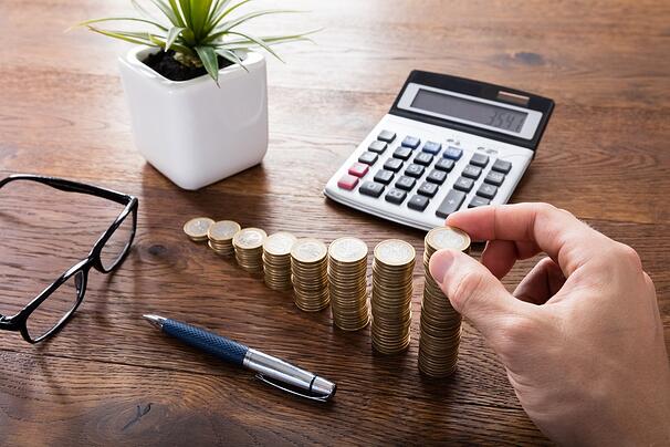 uma mão empilhando moedas em uma mesa de madeira. há uma calculadora e um vaso na mesa, em referência a o que é tesouro ipca