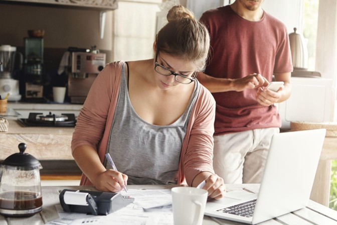 Mulher jovem de óculos concentrada organizando contas da casa com calculadora e notebook na mesa da cozinha, aplicando técnicas de economia doméstica.