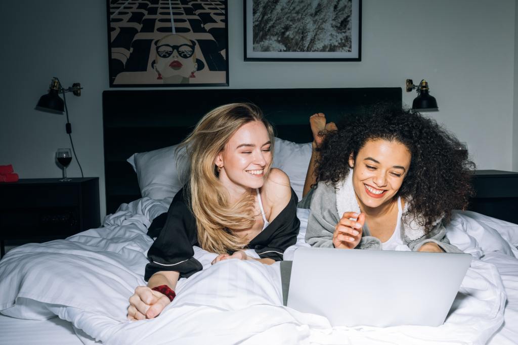 A primeira imagem é uma foto de duas mulheres deitas na cama, com um notebook aprendendo sobre Open Investment coma a Rico.