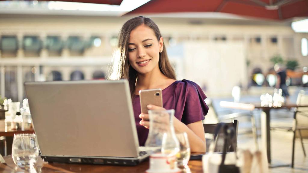 A imagem mostra uma mulher segurado um celular e utilizando o notebook em uma mesa em referência à busca por entender quem está isento de imposto de renda.