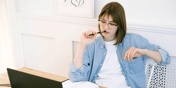Mulher sentada em uma cadeira com a mão segurando uma caneta apoiada em sua boca e uma mesa à sua frente com um notebook e um caderno aberto.