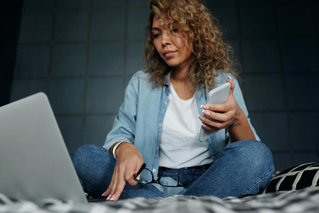 Mulher sentada na cama em frente ao computador com um celular na mão aprendendo sobre Swing trade com a Rico.