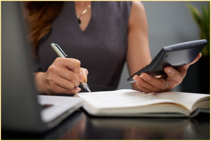 Mulher usando calculadora e caneta para anotar em um caderno como fazer uma reserva de emergência financeira ao lado de um notebook.