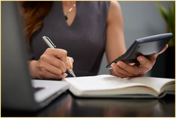 Mulher usando calculadora e caneta para anotar em um caderno como fazer uma reserva de emergência financeira ao lado de um notebook.