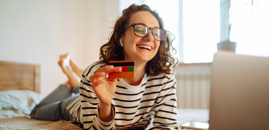 Mulher sorridente deitada em sua cama de forma descontraída enquanto sorri e segura seu cartão de crédito Rico ao encontrar uma promoção de Black Friday em seu notebook.