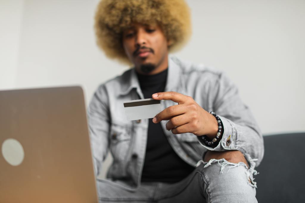 homem, em frente a um notebook, segurando um cartão de crédito.