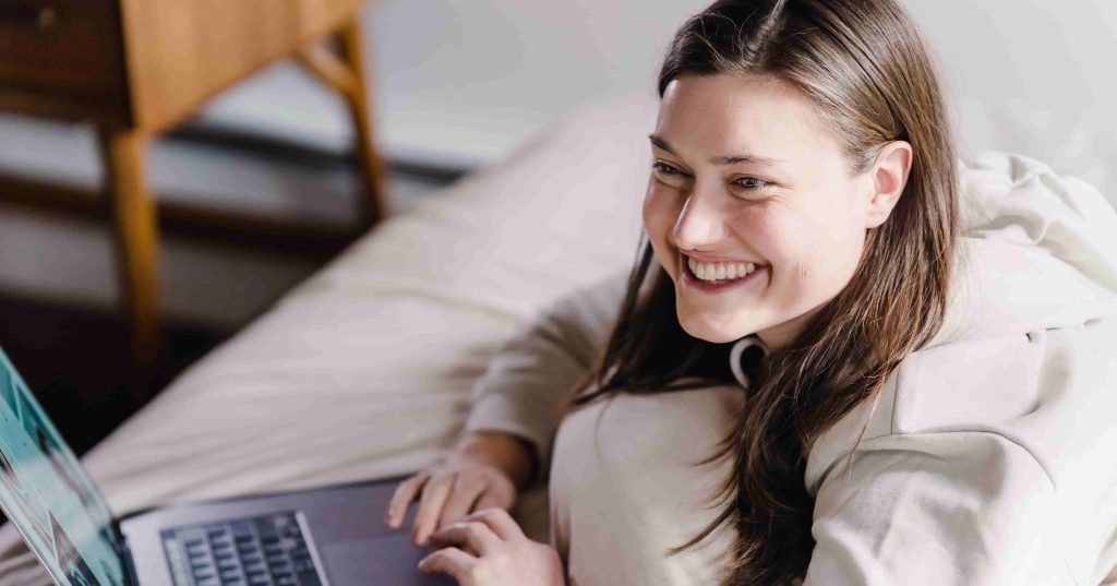 a imagem mostra uma mulher deitada em uma cama, utilizando um notebook, para pesquisar sobre a plataforma trader Profit, e sorrindo.