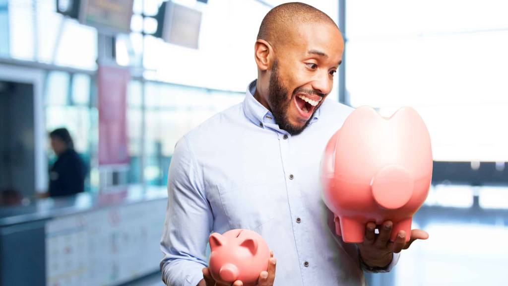 Um homem segura dois cofres no formato de porquinho cor de rosa: um menor na mão direita e o maior na esquerda. Ele está feliz olhando para o cofre maior, considerando o bom desempenho dos seus investimentos de curto prazo.