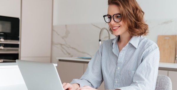 Mulher jovem e sorridente usando óculos e camisa listrada, sentada em uma cozinha moderna enquanto trabalha no notebook.