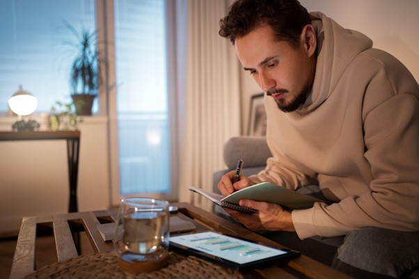 Homem sentado em um sofá fazendo anotações em um caderno ao olhar para um tablet em cima de uma mesa à sua frente.