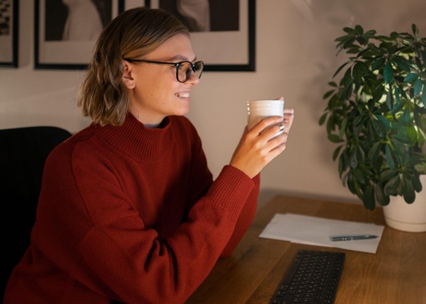 Mulher sentada com os cotovelos apoiados em uma mesa, segurando uma xícara de café e sorrindo, com um teclado à sua frente.