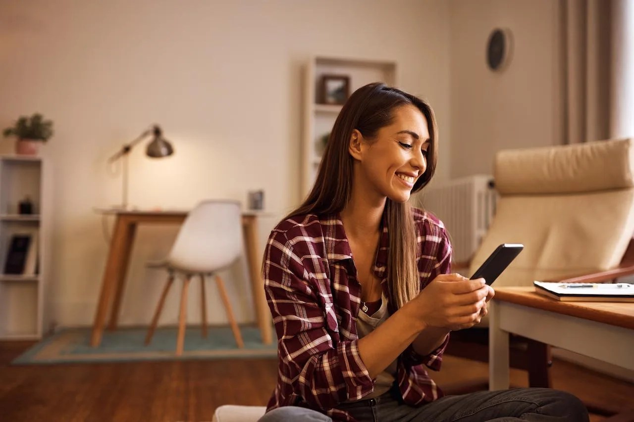 Mulher sentada em uma sala de estar, sorridente enquanto  utiliza a nova funcionalidade "Meus objetivos" da Rico.