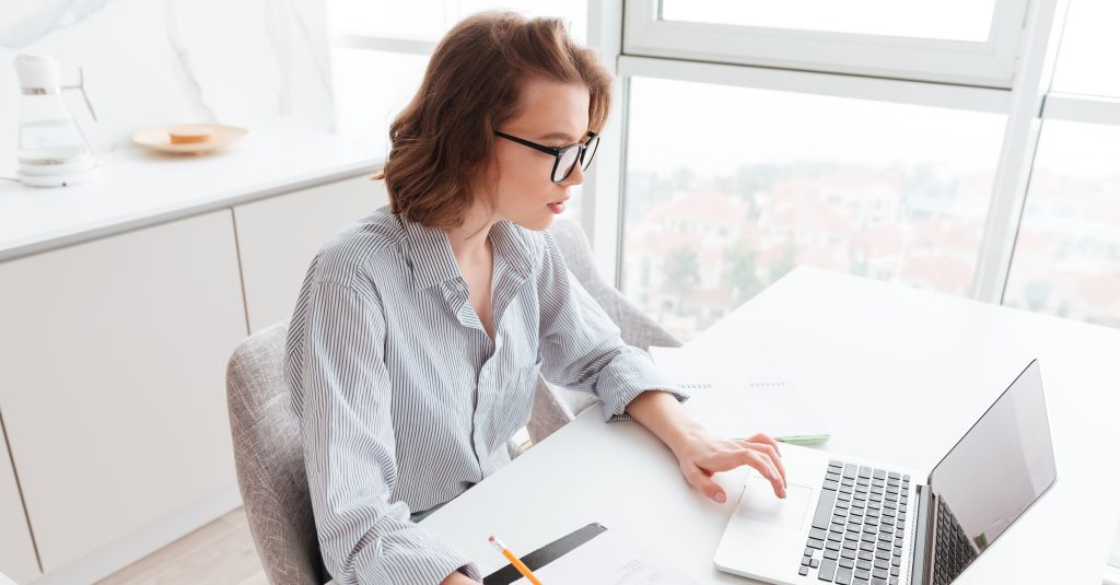 Mulher branca sentada à mesa, pesquisando no notebook sobre o que é IGP-M e seu impacto no aluguel, enquanto faz cálculos com lápis em um papel com valores do IGP-M acumulado.