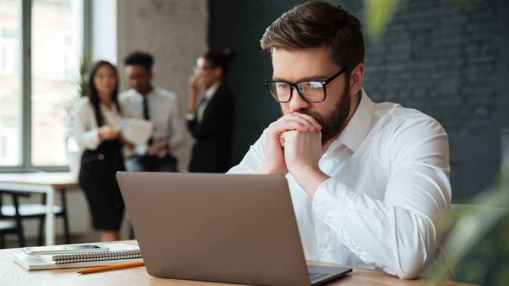 Na imagem, vemos um homem concentrado analisando dados em seu laptop, possivelmente avaliando as tendências e previsões do mercado para tomar decisões sobre investimentos em contratos futuros de Bitcoin. Ele está em um ambiente de escritório moderno, onde outros colegas estão engajados em conversas ao fundo.