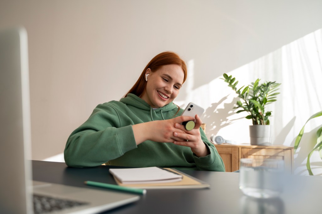 Imagem mostra uma mulher sorrindo e segurando o celular com as duas mãos, apoiada em uma mesa, com papéis e um notebook em frente a ela, e conferindo os pontos no cartão da Rico.