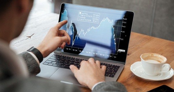 A imagem mostra um homem usando um notebook em cima de uma mesa e apontando com uma das mãos para um gráfico que mostra a alta de investimentos internacionais na tela. Ao lado do notebook, está uma xícara de café e um celular.